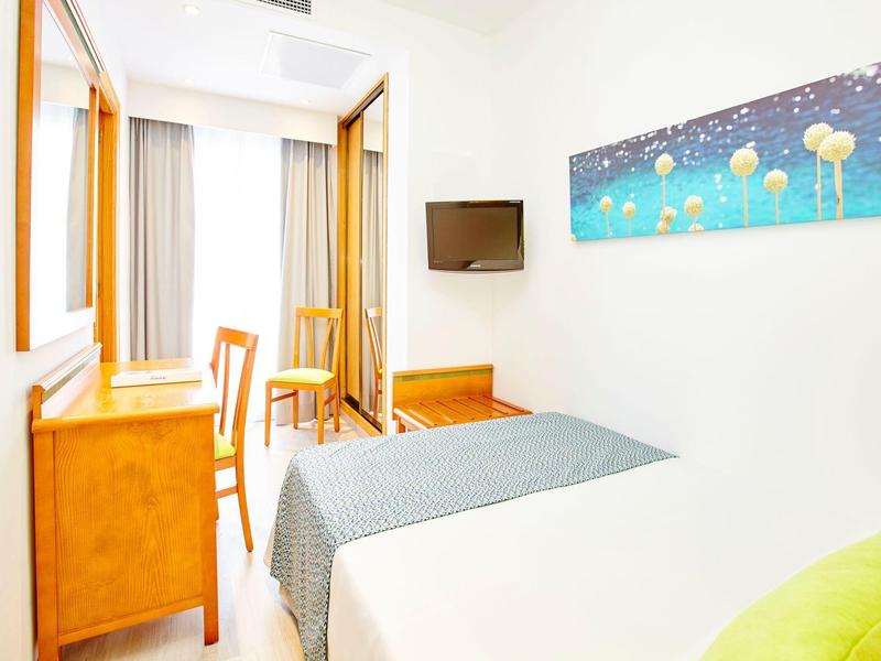 Helles Schlafzimmer mit Bett, grauer Bettdecke, Holzschreibtisch, Stuhl, Wandbild und Wandfernseher.