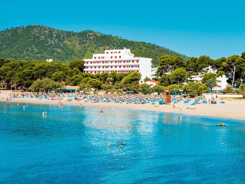 Strand mit türkisblauem Wasser, Sonne, Menschen, weißen Gebäuden und grünen Hügeln im Hintergrund.