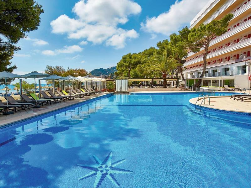 Großer blauer Pool mit Sternmuster, umgeben von Liegestühlen, Bäumen und mehrstöckigem Gebäude.