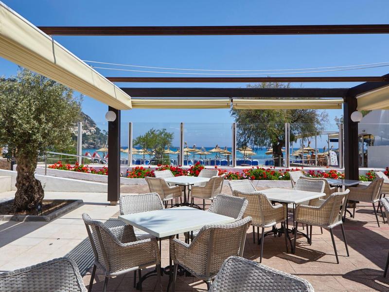 Terrassenbereich mit Tischen und Stühlen, Olivenbäume, blaue See im Hintergrund, sonniger Tag.