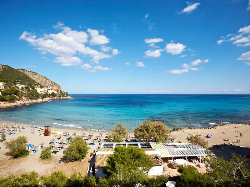 Strand mit blauem Meer, bewölktem Himmel, Sand, Sonnenschirmen und Gebäuden am Ufer.