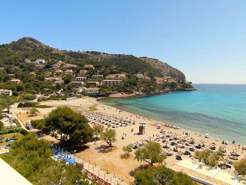 Strand mit Sonnenschirmen, blauem Meer und Hügeln mit Häusern im Hintergrund bei klarem Himmel.