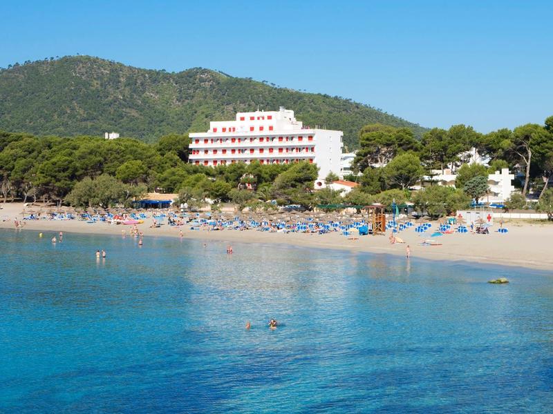 Klarer blauer Meerblick auf einen Sandstrand mit Sonnenschirmen, Bäumen und einem weißen Hotel vor Bergen.
