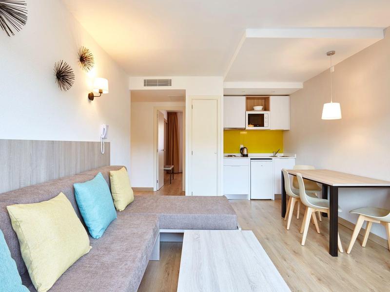 Helles, modernes Wohnzimmer mit grauer Couch, bunten Kissen, kleinem Tisch und offener Küche mit Gelb-Akzenten.