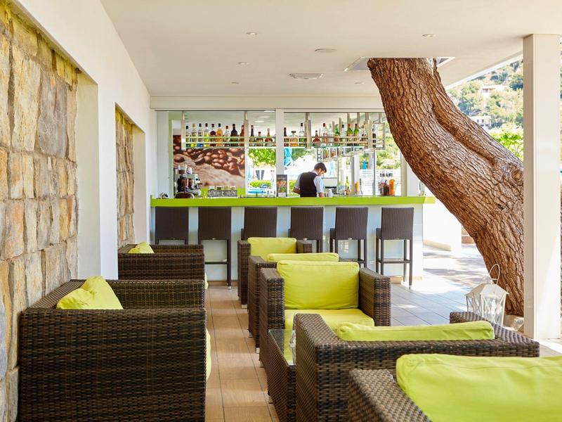 Moderner Außenbereich mit Rattanmöbeln, grünen Sitzkissen und Bar im Hintergrund unter Dach.