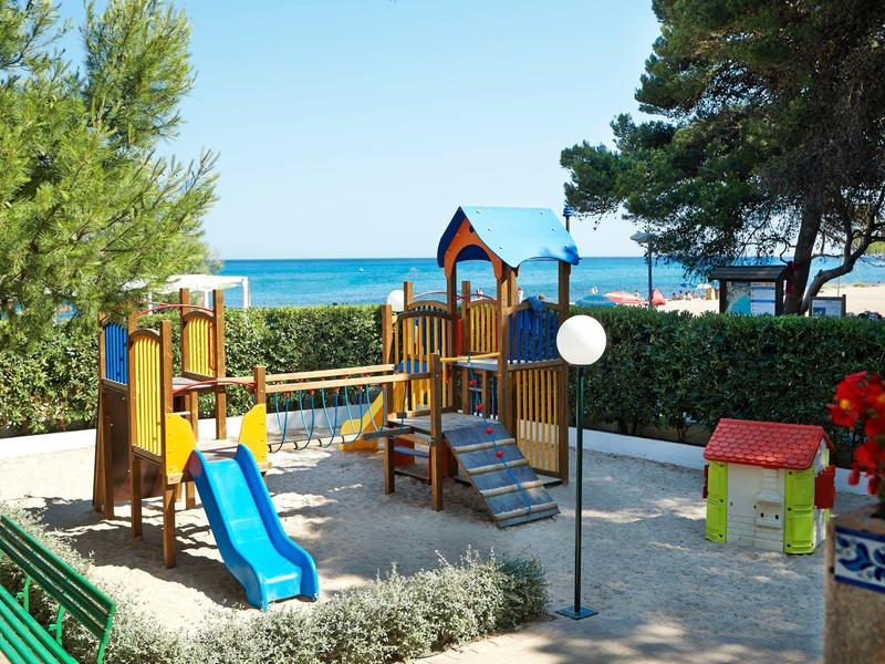 Kinderspielplatz mit Rutsche, Klettergerüst und Haus, umgeben von Bäumen und Meer im Hintergrund.