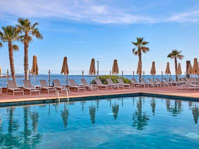 Widok na basen z leżakami i palmami przy morzu pod błękitnym niebem.