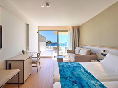 Jasny pokój hotelowy z łóżkiem podwójnym, sofą, biurkiem i widokiem na taras z widokiem na morze.