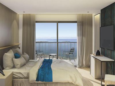 Modern hotelkamer met zeezicht, groot bed, tv en balkon met stoelen.