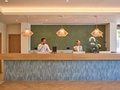 Recepcja hotelowa z dwoma pracownikami za nowoczesnym stanowiskiem i trzema lampami wiszącymi.