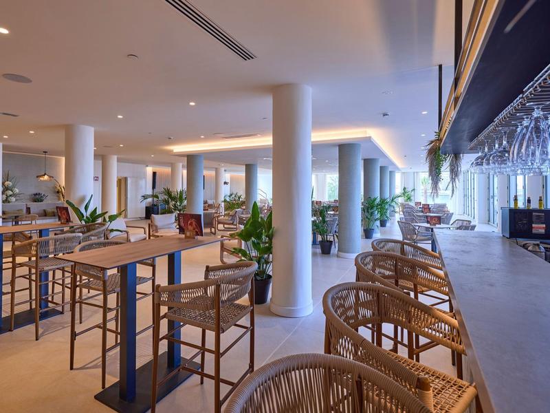 Nowoczesna restauracja z wysokimi stolikami, krzesłami i barem w jasnej, otwartej przestrzeni.