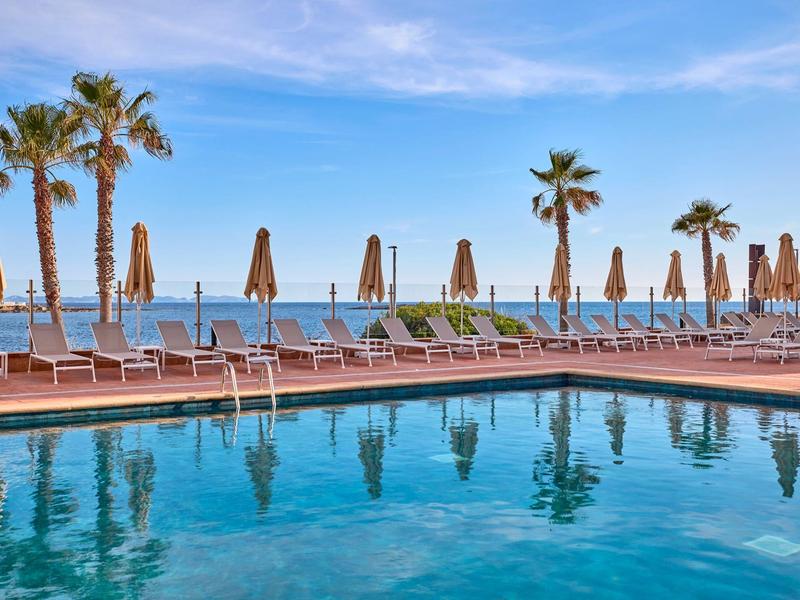 Widok na basen z leżakami i palmami przy morzu pod błękitnym niebem.