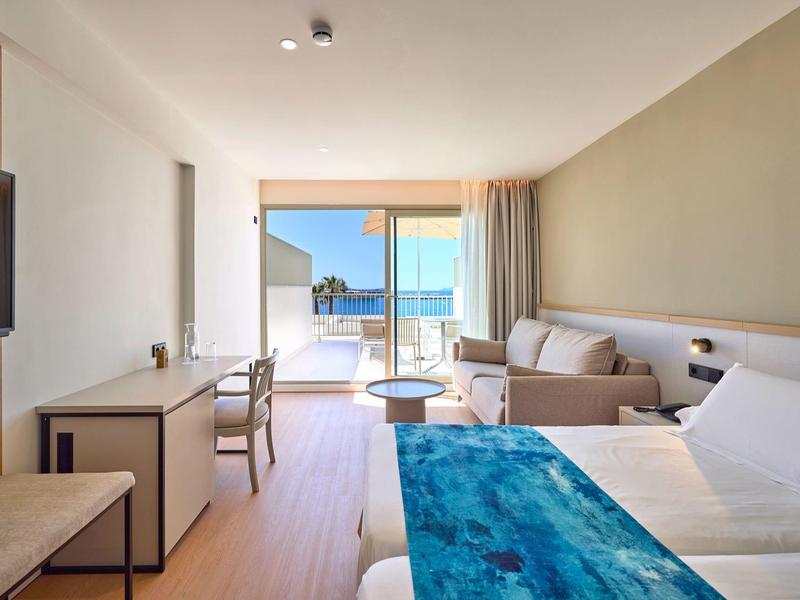 Jasny pokój hotelowy z łóżkiem podwójnym, sofą, biurkiem i widokiem na taras z widokiem na morze.