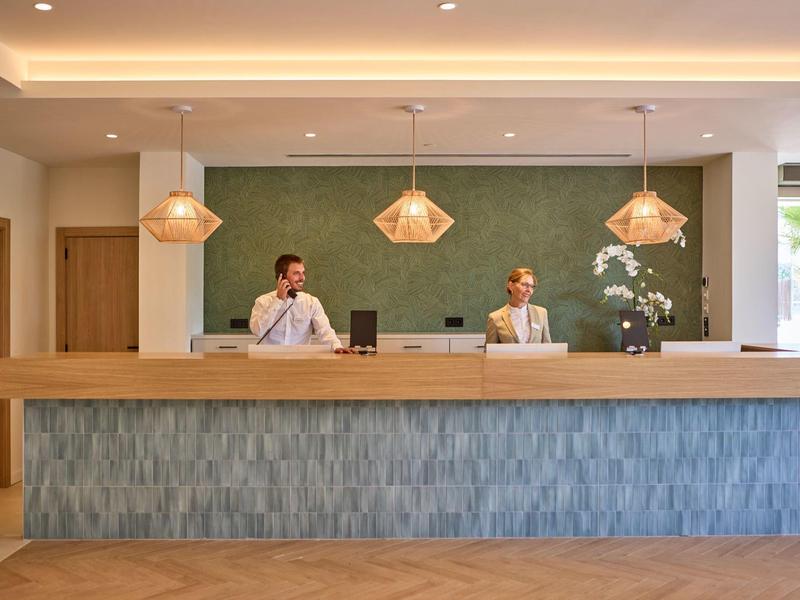 Recepcja hotelowa z dwoma pracownikami za nowoczesnym stanowiskiem i trzema lampami wiszącymi.