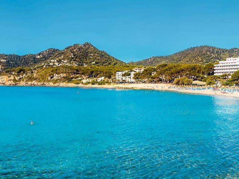 Široká písečná pláž s čistou modrou vodou, obklopená hotely a kopci.