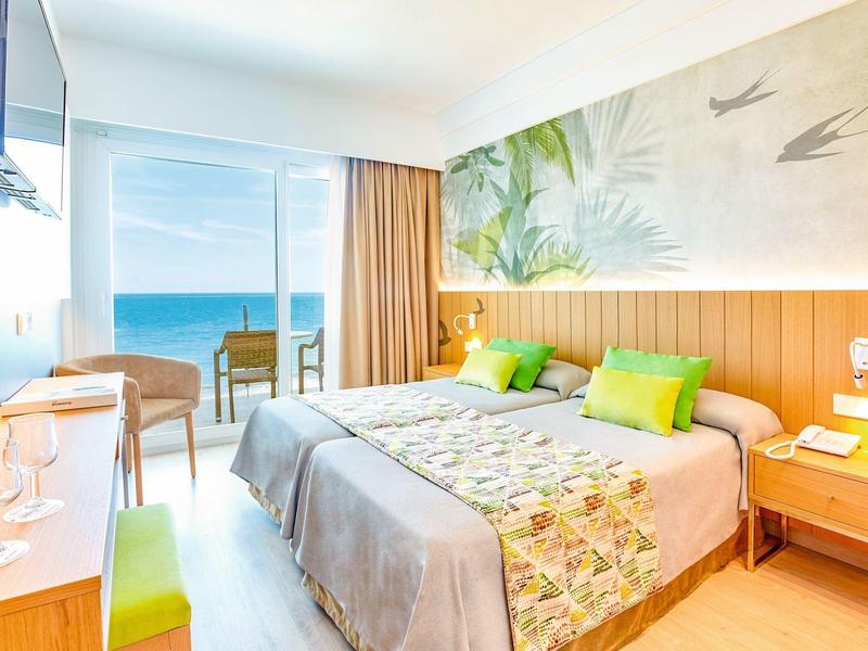 Světly hotelový pokoj s výhledem na moře, manželskou postelí a moderním interiérem.