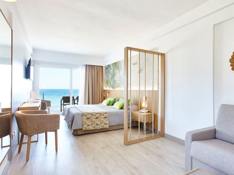 Moderní hotelový pokoj s manželskou postelí, posezením a výhledem na moře přes velká okna.