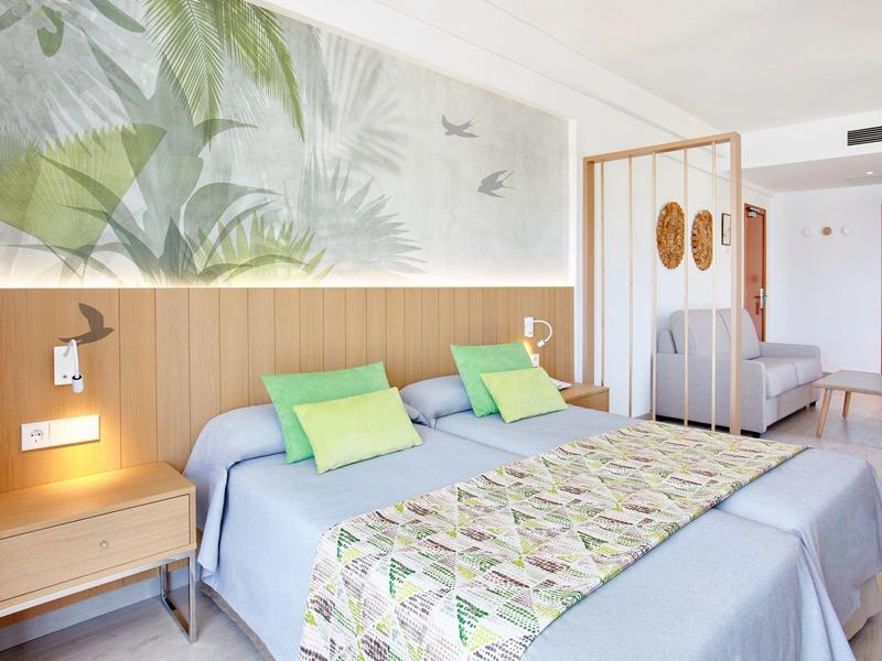 Světý hotelový pokoj se dvěma postelemi, zelenými polštáři a přírodní nástěnnou dekorací.