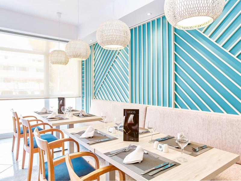 Světlá restaurace s modrými stěnami, dřevěnými stoly a židlemi s modrými polštáři.