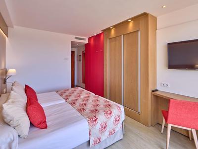 Nowoczesny pokój hotelowy z podwójnym łóżkiem, czerwonym akcentem, szafą i telewizorem ściennym.