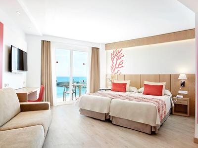 Jasny pokój hotelowy z dwoma łóżkami pojedynczymi, sofą, biurkiem i balkonem z widokiem na morze.