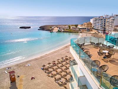 Vista de una playa soleada con tumbonas y un balcón de hotel adyacente con mesas.