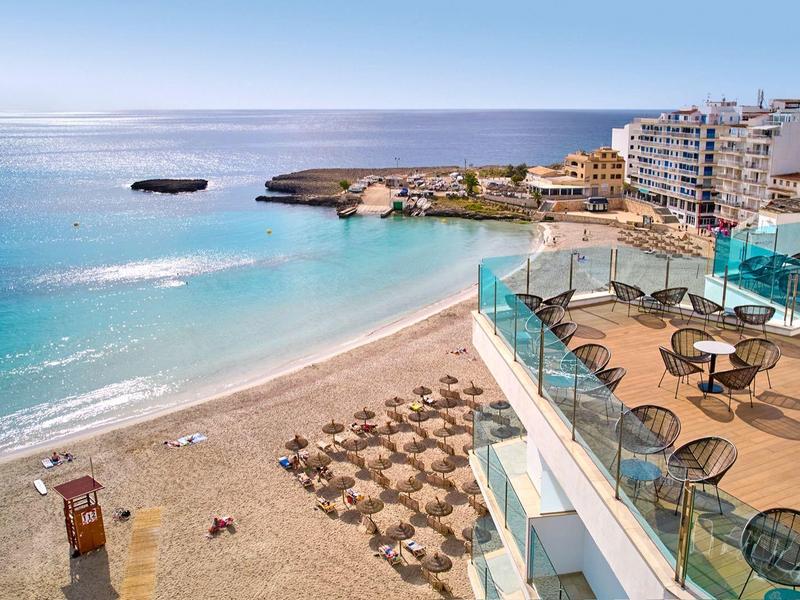 Widok na słoneczną plażę z leżakami i hotel z tarasami nad morzem.