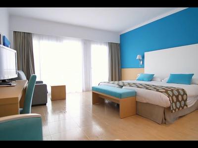 Lichte hotelkamer met een tweepersoonsbed, blauwe accentmuur, bureau en tv bij grote ramen.