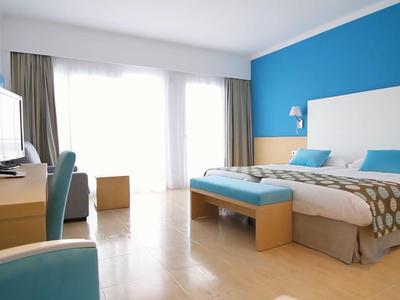 Lichte hotelkamer met twee eenpersoonsbedden, blauwe accentmuur, bureau en groot raam met gordijnen.