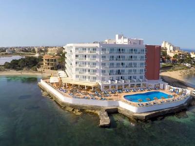 Wit hotel met zwembad op rotsachtige kustlijn omgeven door helderblauwe zee bij daglicht.