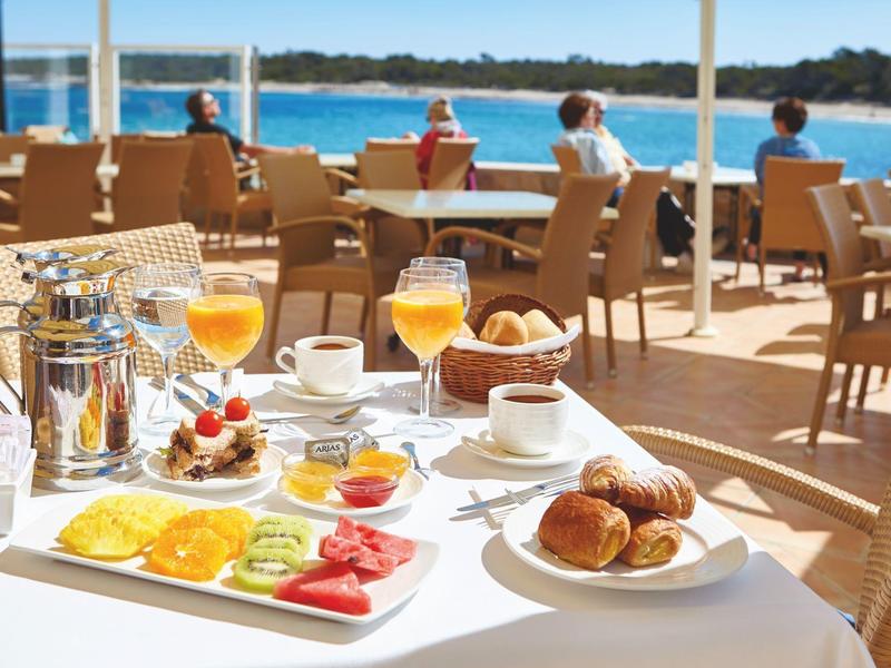 Ontbijt buiten met sinaasappelsap, croissants, fruit en zeezicht.
