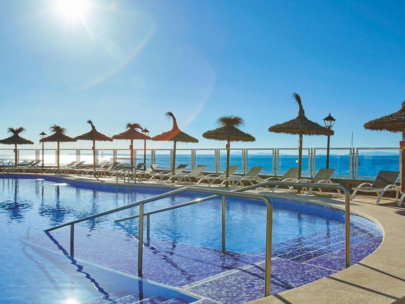 Buiten zwembad met rieten parasols en ligstoelen aan zee onder een heldere blauwe lucht.