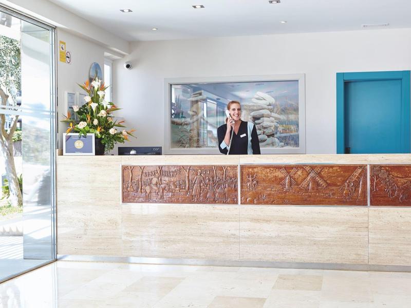 Hotel lobby met receptionist, houten balie, blauwe deur en groot raam met uitzicht op buitenbomen.