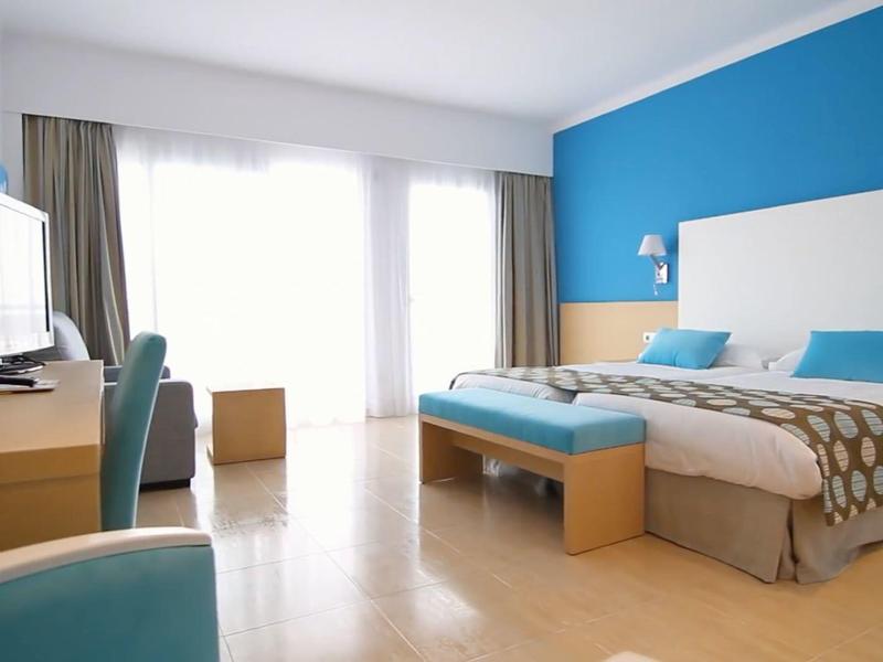 Lichte hotelkamer met twee eenpersoonsbedden, blauwe accentmuur, bureau en groot raam met gordijnen.