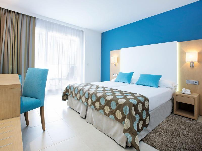 Lichte hotelkamer met blauwe accentmuur, tweepersoonsbed, houten meubels en groot raam met gordijnen.