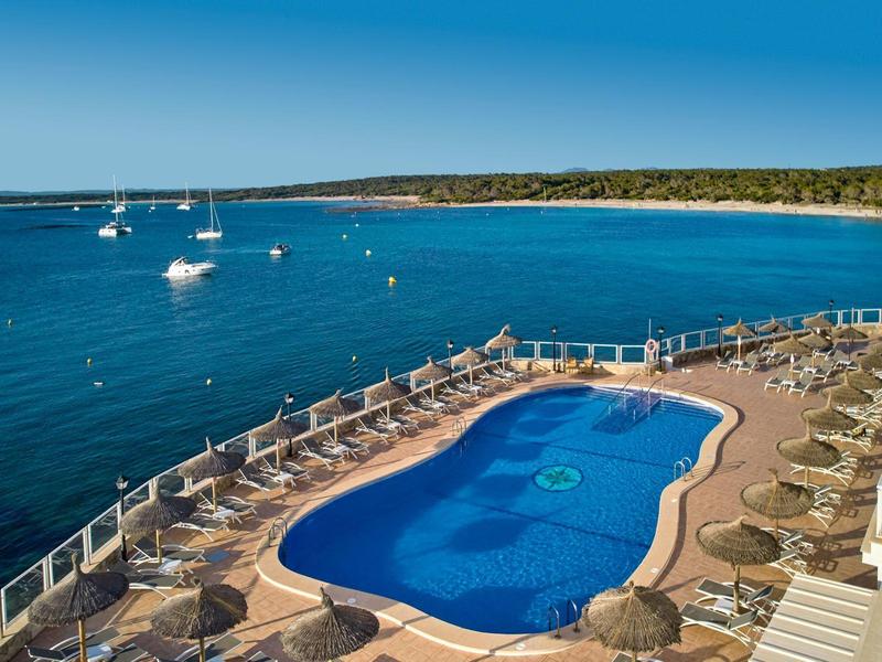 Buitenzwembad met ligstoelen met uitzicht op een rustige blauwe zee en zeilboten onder een heldere lucht.