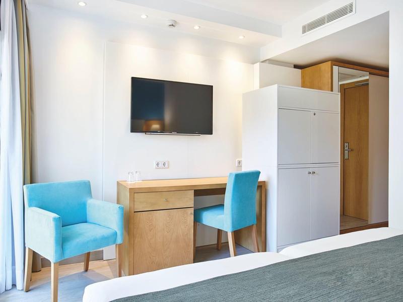 Hotel room with blue chairs, wooden desk, TV on white wall, and refrigerator near wooden door.