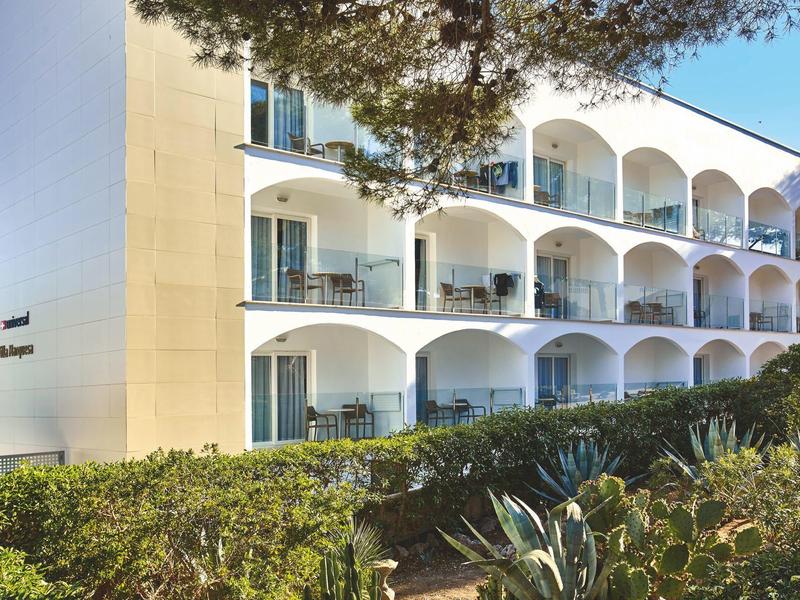 Three-story hotel with arches on balconies surrounded by greenery under a clear sky.