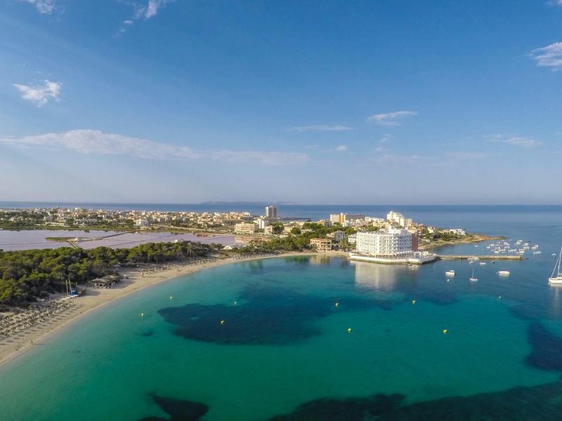 Coastal resort with clear turquoise waters, white sandy beach, and buildings under a blue sky.