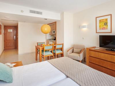 Camera d'hotel luminosa con letto matrimoniale, tavolo da pranzo, poltrona e TV su una cassettiera.