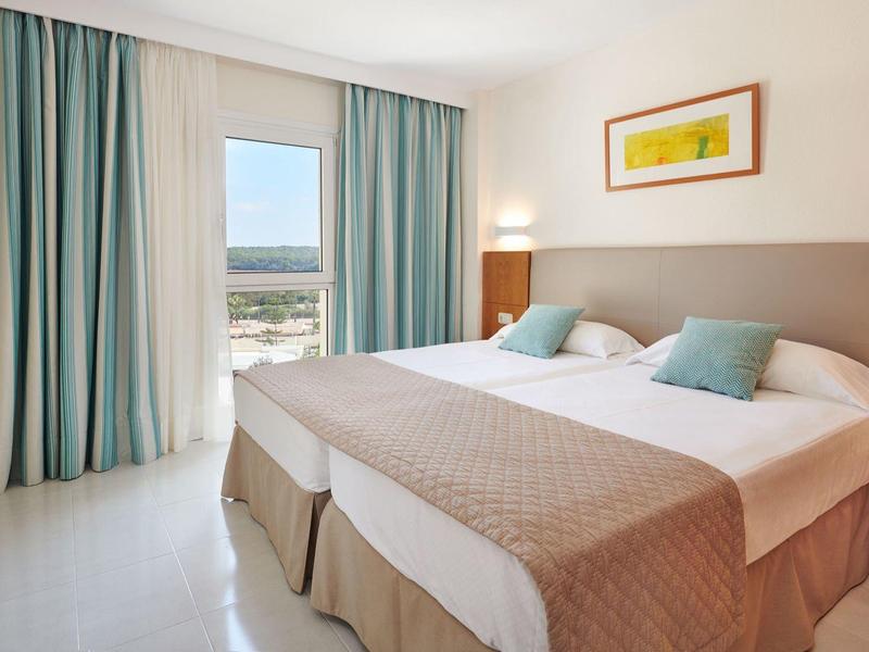 Camera d'hotel luminosa con due letti, tende turchesi e vista sulla piscina.