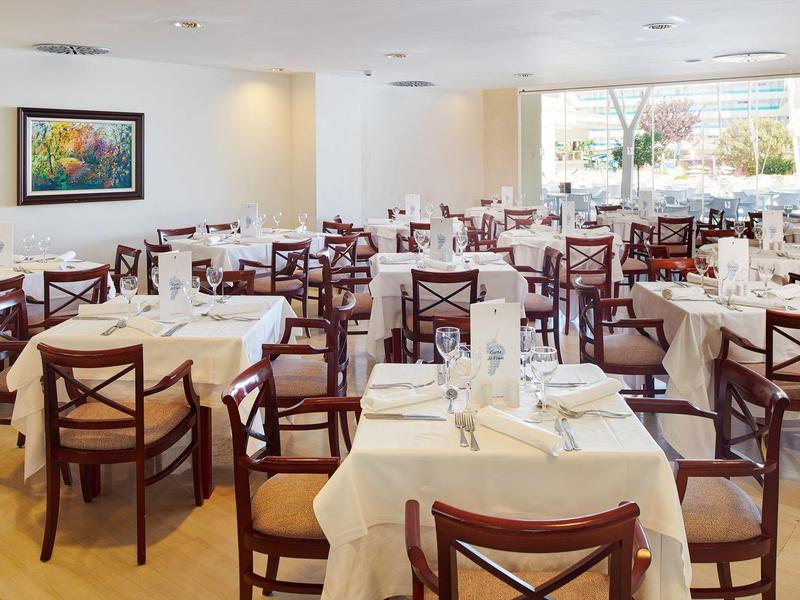 Ampia sala da pranzo luminosa con molti tavoli e sedie, apparecchiata con tovaglie bianche.