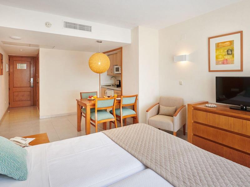 Camera d'hotel luminosa con letto matrimoniale, tavolo da pranzo, poltrona e TV su una cassettiera.