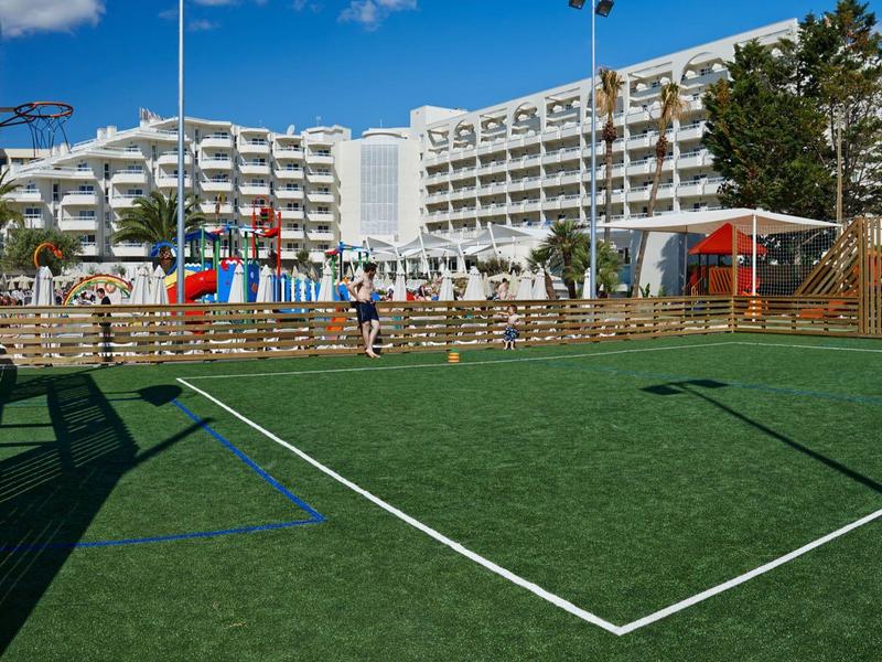 Campo sportivo moderno con erba sintetica davanti a un grande hotel con balconi bianchi.