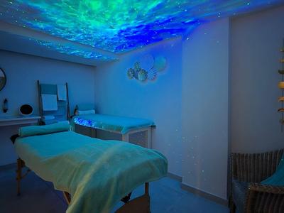 Przyciemniony pokój relaksacyjny z łóżkiem do masażu, niebieskim oświetleniem i spokojną atmosferą.