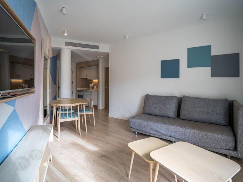 Nowoczesny pokój hotelowy z sofą, stolikami oraz częścią jadalną w jasnym wnętrzu.