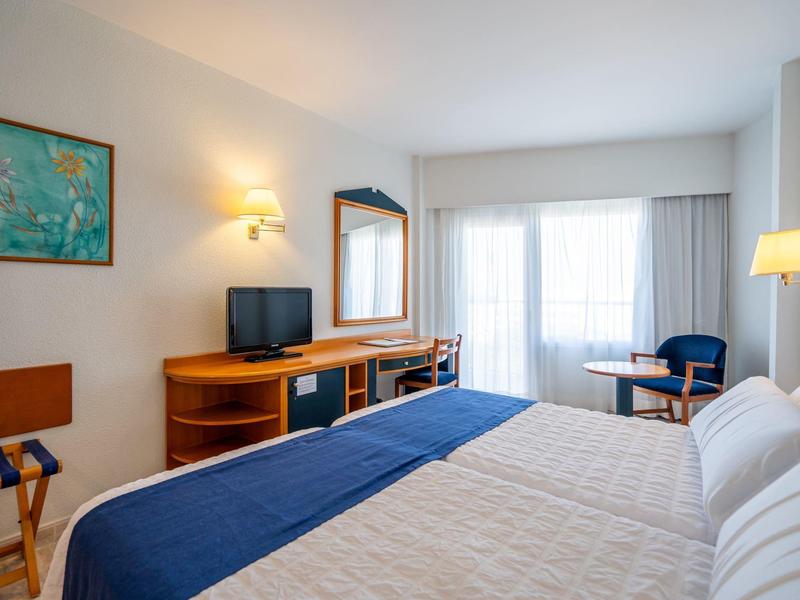 Jasny pokój hotelowy z dwoma łóżkami, telewizorem, fotelem i dużym oknem wpuszczającym światło dzienne.