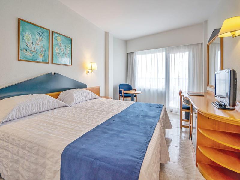 Jasny pokój hotelowy z podwójnym łóżkiem, niebieskimi akcentami, dwoma obrazami i dużym oknem.