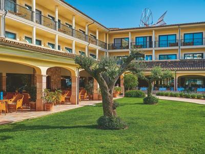 Edificio del hotel con patio, césped verde y olivos bajo cielo azul