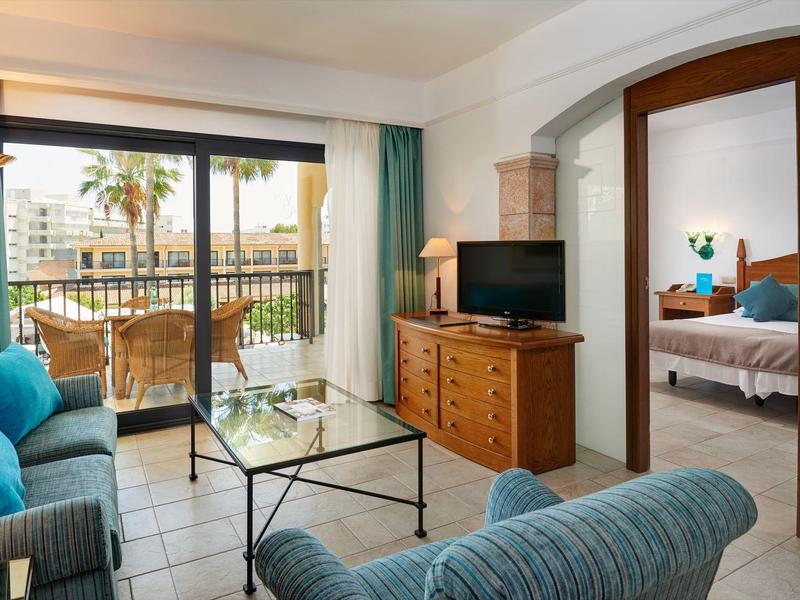 Gezellige hotelkamer met zithoek, glazen tafel, tv en balkon met uitzicht op palmbomen.